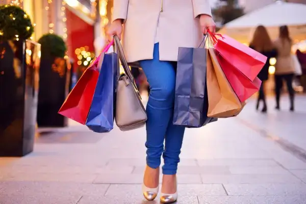 Mulher caminhando com várias sacolas de compras, sem mostrar o rosto, representando decisões de consumo e comportamento financeiro.