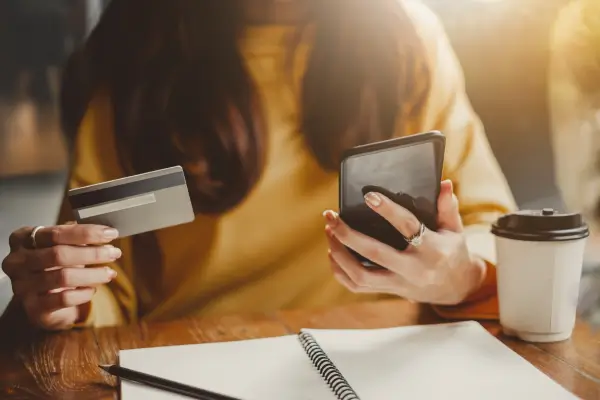 Mulher segura um celular em uma mão e um cartão de crédito na outra, com um caderno de anotações sobre a mesa — representação de hábitos de consumo e tomada de decisão financeira.